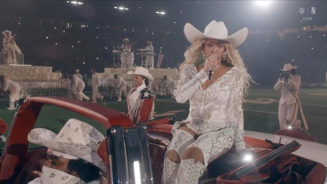 A stage performance featuring a female artist in a sparkling white outfit and cowboy hat (Beyonce), singing into a microphone while sitting on a red convertible. Accompanying her are dancers in white outfits and a musician in the background. The setting appears to be a large
