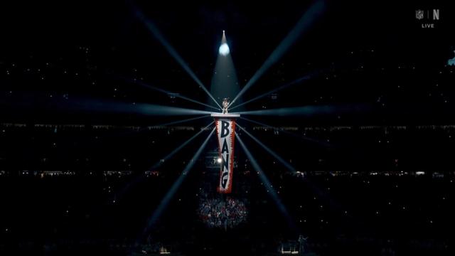 A performer (Beyonce) stands atop a large banner reading "BANG," surrounded by dramatic stage lights radiating from above in a dark arena filled with spectators.