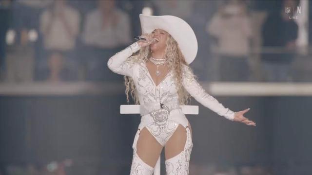 A performer in a white, sparkling outfit and large white cowboy hat (Beyonce) sings passionately on stage, with a microphone in hand. The audience is blurred in the background, creating a dynamic concert atmosphere.