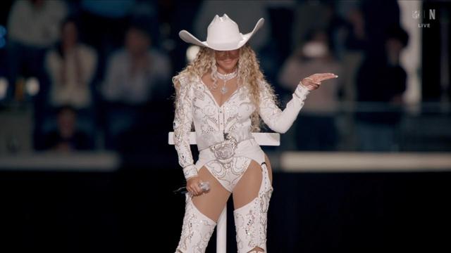 A performer in a sparkling white outfit and a wide-brimmed white cowboy hat (Beyonce) gestures on stage, with a microphone in hand. The audience is blurred in the background.