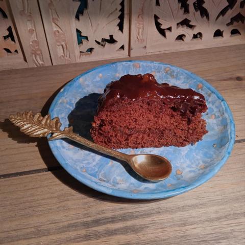 A slice of rich brown chocolate sponge cake sits on a round blue pottery plate, on a wooden surface  A leaf shaped brass spoon sits diagonally across the plate. Behind is a cut Wisden fretwork door.