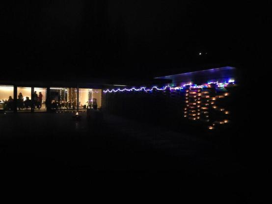 Ein Foto, aufgenommen bei Nacht, zeigt ein Gebäude mit großen Fenstern. Im Inneren des Gebäudes sitzen und stehen Personen an Tischen, und es gibt Lichterketten an der Decke. Vor dem Gebäude steht ein Pfahl mit einer brennenden Flamme. Auf der rechten Seite des Gebäudes befindet sich eine Mauer. Auf dieser Mauer sind Kerzen in einem Muster angeordnet, welches RRS, die Initialen von Rechtsruck Stoppen ergibt und es gibt eine weitere Lichterkette entlang der Oberseite der Mauer. Der Himmel ist dunkel, und ein kleiner heller Punkt, wahrscheinlich der Mond, ist oben rechts sichtbar.