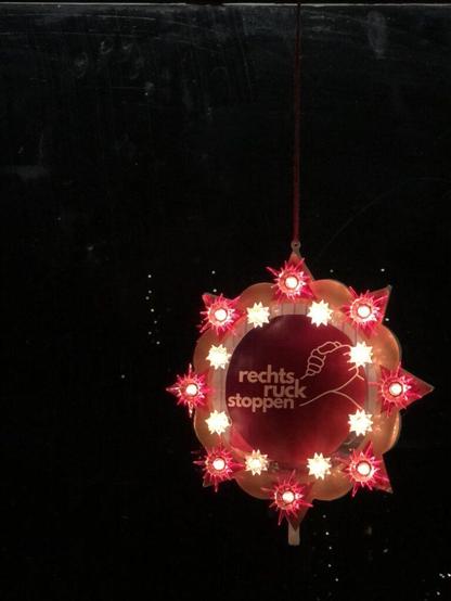 Ein alter Holztisch ist mit verschiedenen Gegenständen dekoriert. Auf dem Tisch liegen mehrere Kerzen in kleinen Haltern, die brennen und Licht spenden. Es gibt verschiedene Früchte wie Orangen und Äpfel, teilweise mit grünen Blättern. Zwei Flyer liegen auf dem Tisch. Auf dem vorderen Flyer steht „Rechtsruck stoppen!“. Ein stilisiertes Bild zeigt zwei Hände, die sich festhalten. Der Hintergrund des Flyers ist rot und lila. Im Hintergrund sieht man Lichterketten, die für eine gemütliche Atmosphäre sorgen. Hinten stehen Stühle und eine Tasche liegt auf einem der Stühle. Der Raum wirkt dunkel, aber durch die Kerzen und Lichterketten wird es hell und warm.