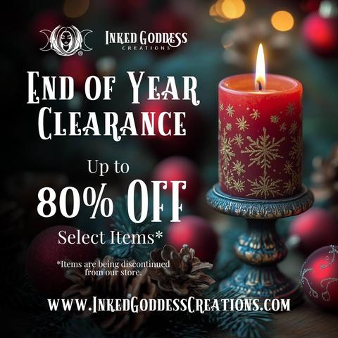 A red pillar candle is burning on an antiqued metal stand on a table. The candle is lit and has gold snowflakes dotting its surface. The text reads, "End of Year Clearance; Up to 80% off Select Items*; *Items are being discontinued from our store." Presented by Inked Goddess Creations.