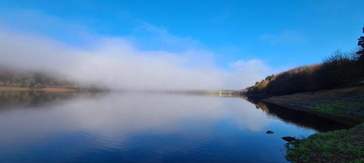 A photo of mist/a low cloud inversion over a large reservoir. The water is so flat that it's perfectly reflecting the blue sky above, and the mist/cloud that is barely above the water. The bright sky and cloud is contrasted with a dark, curving bank on the right of the image.