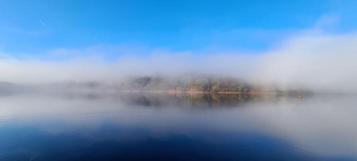 Another photo of the reservoir in the previous image. Again, the water reflects the bright blue sky above and the white mist just above the water. The tree covered far bank is visible through a break in the mist. With the reflection in the water, the image looks almost like it's mirrored top/bottom, and left/right.