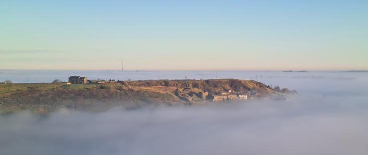 Yet another cloud inversion picture, again showing the mist in the valleys around hills. This is long distance shot, showing the mist extending in the valleys for miles. A few hilltops peak out through the mist, including one on the mid-ground. In the very far distance, a large TV antenna (Emley Moor mast) is just about visible, as is a much small tower (a Victorian folly tower on Castle Hill in Huddersfield).