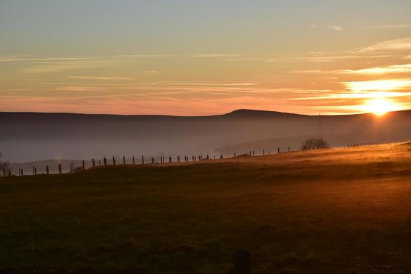 A photo of a valley, filled with fog, the sun is setting behind a hill and there is a rickety wire fence across the middle.