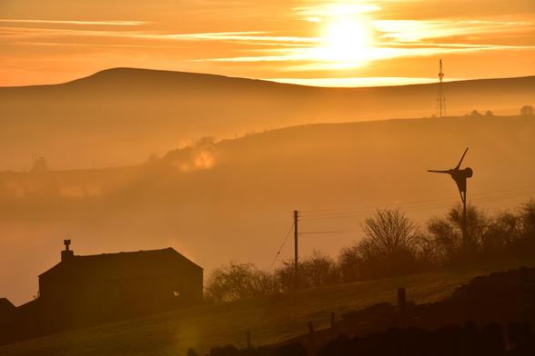 A sun starting to set behind a hill. There is a silhouette of an old farm house and a wind turbine in the foreground. It is very misty.