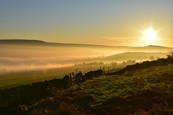 A valley filled with fog. There is a yorkshire stone wall in the foreground and a sun is starting to set behind the hill.