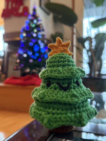 a green crochet amigurumi tree named “douglas the fir”, with beady black eyes and a short smile, brown stubby trunk, and a yellow star topper. in the background is my parents’ christmas tree in mostly blue lights near a fireplace, and a large bird of paradise plant. 