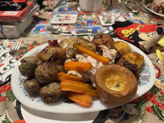 A plate of festive food featuring roasted turkey; kilted sausages; pork, sage & onion stuffing; carrots, Yorkshire pudding and roast potatoes, all set on a decorated table with Christmas-themed items and a pair of Mickey Mouse socks.