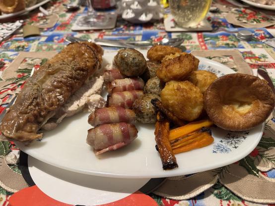 A plate of festive food featuring roasted turkey; kilted sausages; pork, sage & onion stuffing; carrots, Yorkshire pudding and roast potatoes, all set on a decorated table.