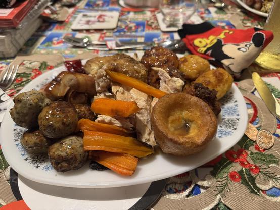 A plate of festive food featuring roasted turkey; kilted sausages; pork, sage & onion stuffing; carrots, Yorkshire pudding and roast potatoes, all set on a decorated table with Christmas-themed items and a pair of Mickey Mouse socks. Topped with gravy. 