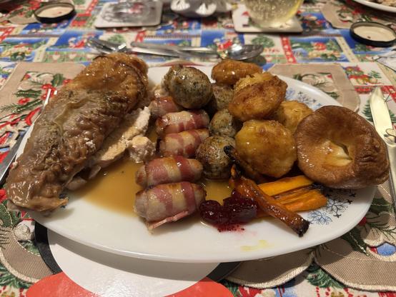 A plate of festive food featuring roasted turkey; kilted sausages; pork, sage & onion stuffing; carrots, Yorkshire pudding and roast potatoes, all set on a decorated table. Topped with gravy. 