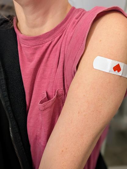 White woman wearing a pink tee shirt, exposing her left arm with a white CVS band aid on her arm that was just vaccinated. 