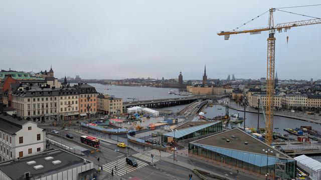 Big intersection with pedestrian crossings, new halls with glass roofs and entries to the metro, historical buildings on the left, areas with construction machined in between, a construction crane behind it, Gamla Stan in rhe background