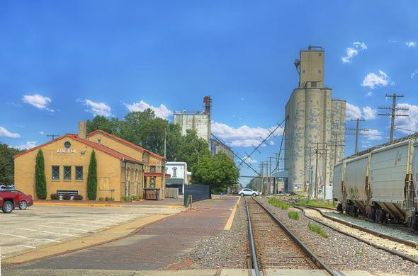 A railway track runs through a small town scene, with a brick building labeled "Abilene" on one side and large industrial silos on the other.