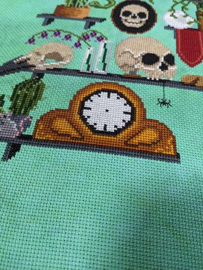A potion of a cross stitch piece showing a wooden mantle clock with not hands and a spider hanging above it.
