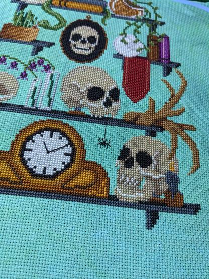 Portion of a cross stitch piece showing a mantle clock reading 11:10. Next to it is a human skull facing slightly to the left with a candle to the right front.