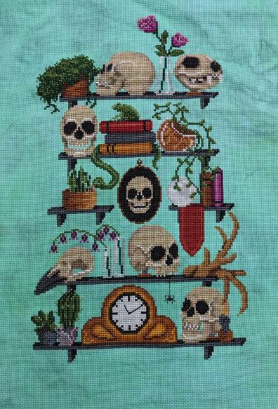 Completed stitching on cross stitch showing five levels of shelves with a variety of skulls, plants, and books. There is also a fun little frog, snake, and spider hanging out.