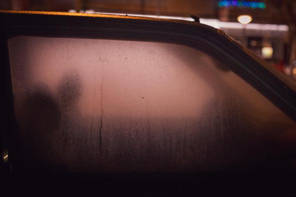 Fotografía donde se ve una ventanilla empañada de un coche desde fuera. Se perciben un par de sombras que son los asientos. Tangencialmente, en la parte superior derecha, se ve algo de las luces de la ciudad desenfocadas (es de noche).