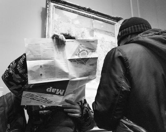 Black and white photo of two people mostly obscuring a painting in an art gallery. One, facing the camera, is holding a map of the gallery, apparently upside down. The other person is turned away from the camera, looking down at something… their own map?