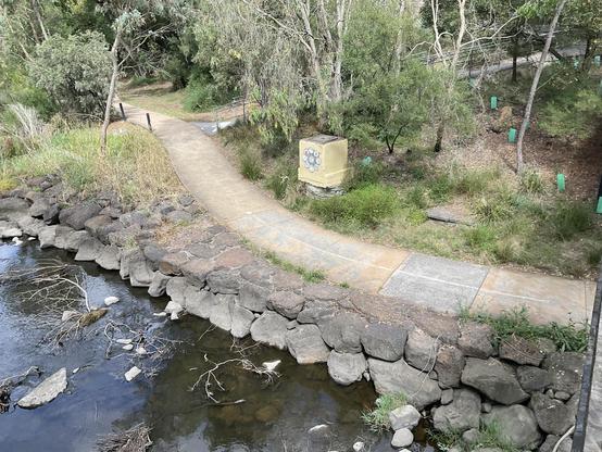 Merri Creek and bike path Scene seen from above. Rock reinforcement of creek edge 
A path also winds up hill to road. 
A yellow painted street guttering storm water drain access  box has a mosaic on it.