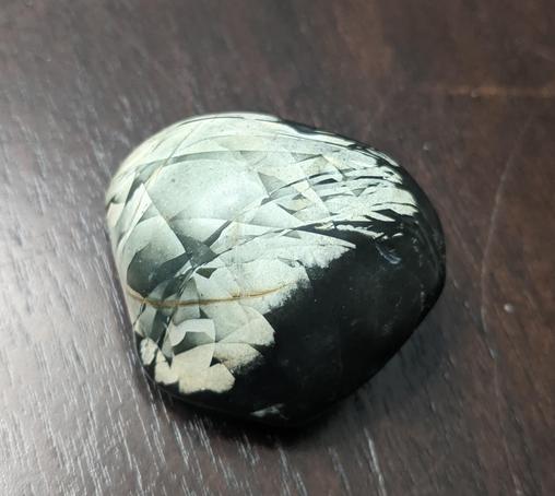 A dark colored rock with overlapping triangles of white and pale green sitting on a dark wood table. No idea what kind of rock except cooooool.