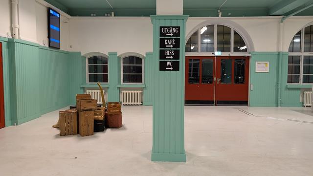 Big waiting hall with man-high wood pannels painted in turquoise, wooden boxes arranged in the middle of the hall, four black signs leading to the utgång, kafé, hiss and wc on a pillar