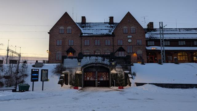 A brick station building with tracks and overhead wires in front, the entrance is located underneath the tracks and built as a stone arch, lettering "Haparanda Järnvägsstation" above the door