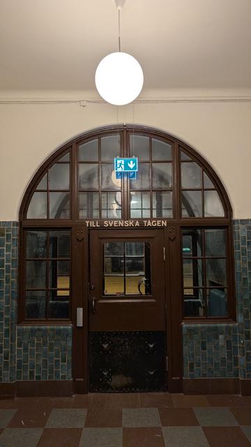 a dark brown wooden door in a arch-shaped opening in the wall, surrounded with glass, lettering "till svenska tågen", blue-green tiles at the wall