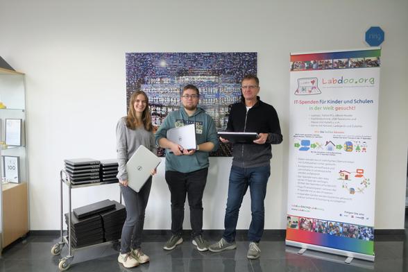 Eine Mitarbeiterin und ein Mitarbeiter von Ferber-Software und Armin vom Labdoo Hub Iserlohn stehen in einem Bürogebäude vor einer weißen Wand mit einem großen Bild, daneben ein Roll-Up von Labdoo. Auf einem Rollwagen liegen mehrere Stapel mit Laptops. Alle Personen halten je einen Laptop in der Hand und schauen in die Kamera.