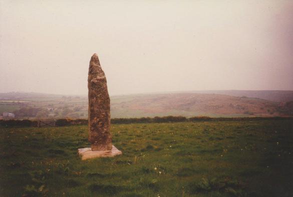 Looking out across the moors of the parish of Wendron from Burras Menhir near Porkellis. The bullet-shaped stone stands left of centre, secured by a diamond shaped base of paler granite. The stones glisten in the rain. The field is green with a low hedge in the middle distance. The moors beyond the hedge are misty and gently curved. The sky is grey with a thick mist.
