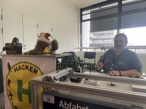 Adelheid sitzt auf dem hohen Tresen und spricht mit einem Menschen hinter dem niedrigen Tresen der dabei ist Fahrgastrechteformulare zu stempeln.