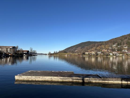 Der Tegernsee, vorne von rechts ins Bild ragt ein Steg. Rechts am Ufer bewaldete Hügel, links ein paar Häuser. Über allem hellblauer Himmel.