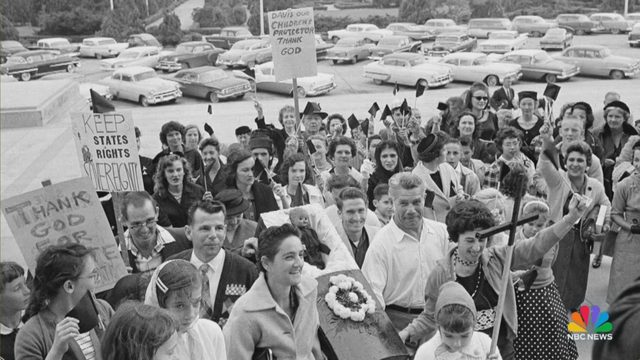 B&W photo of a crowd of white people protesting admitting 5 year old black girl Ruby Bridges into an all-white public school.

Two people can be seen carrying signs touting the "Sovereignty" of "States Rights" while they carry a child-sized coffin with a black baby doll inside.
