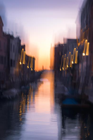 Fotografía vertical donde se ve un paisaje urbano de Venecia, con un canal de agua central. Este paisaje se encuentra difuminado por el movimiento de cámara intencionado. Se puede apreciar una luz del ocaso y como las luces de la ciudad ya están encendidas.