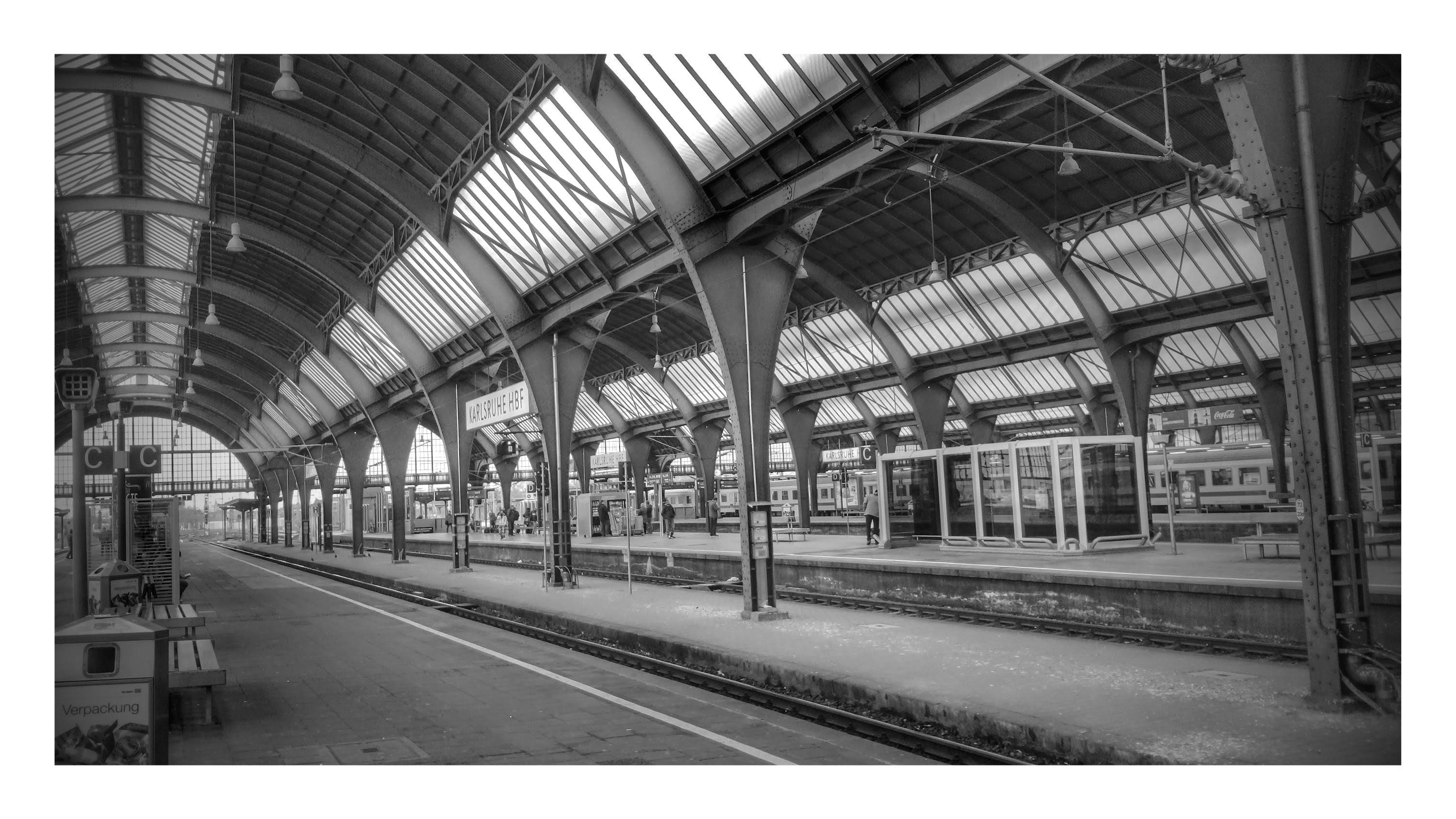 A%20black%20and%20white%20photograph%20of%20a%20mostly%20empty%20train%20station%2C%20indoors%20looking%20down%20an%20empty%20platform%2C%20focusing%20on%20the%20curvature%20of%20the%20roof%20in%20a%20series%20of%20arches%20overhead.%20There%20are%20a%20handful%20of%20people%20on%20the%20next%20platform%20over%2C%20and%20a%20parked%20train%20one%20or%20two%20platforms%20further%20in%20the%20distance.