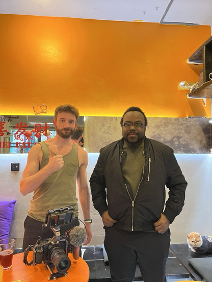 Two men stand behind a table at a cafe. One is wearing a green tank top and giving the camera a thumbs up and the other is wearing a green shirt and black jacket with his hands in his pockets. On the table is a camera a a glass with beer. A cat is sitting on a bench in the background 