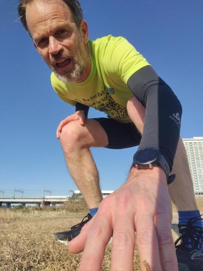 Colin crouched down to take a selfie on his phone after his run.  He looks tired, but strong.  He's wearing a 2025 Osaka marathon t-shirt, black sleeves, black shorts and shoes.