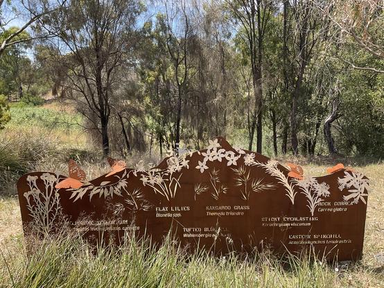 Public art sculpture showing indigenous plants; 
brown rusted iron sheet main panel and butterflies 
With attached silver sheen metal design/ outlines of flowers and grasses. In foreground local green grasses weathered brown by summer sunshine heat.
In background along Creek are stand of indigenous trees with blue sky above. 

Kangaroo Grass (Themeda Triandra)
Flax Lilies (Dianella spp)
Rock Corea (Correa Glabra)
Sticky Everlasting 
Common Everlasting
Melbourne Yellow Gum
Tufted Bluebell (Wahlenbergia)
Eastern Spinebill (Acantharhynchus) 