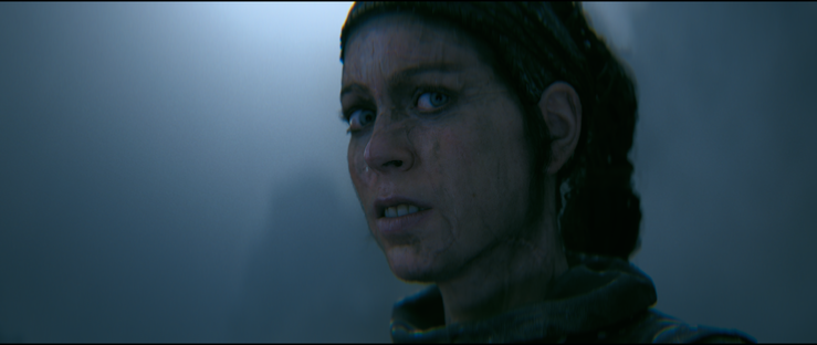 Screenshot from the video game Hellblade 2 showing a close-up portrait of the protagonist Senua, the scene is moody with fog in the background. Senua is looking past the camera, her face dirty and mouth slightly ajar.