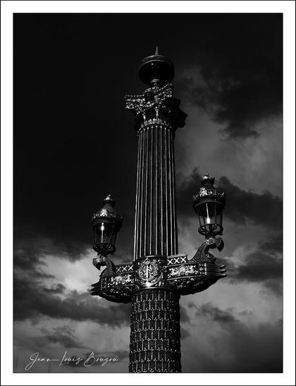 The image is a dramatic black and white photograph of an ornate lamp post against a cloudy sky. The lamp post is tall and features intricate carvings and decorative elements, with three lanterns branching out from the top. The contrast in the image highlights the details of the structure, giving it a striking and imposing appearance.
