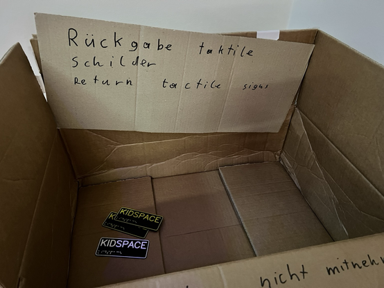 Zu sehen ist ein Pappkarton mit einem Pappschild, auf dem „Rückgabe taktile Schilder - Return tactile signs“ zu lesen ist. In dem Karton liegen drei Schilder mit der Aufschrift „Kidspace“.