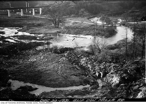 Black and white photo of a muddy riverbank, with a few paths, a long railway bridge, and industrial smokestacks in the distance.