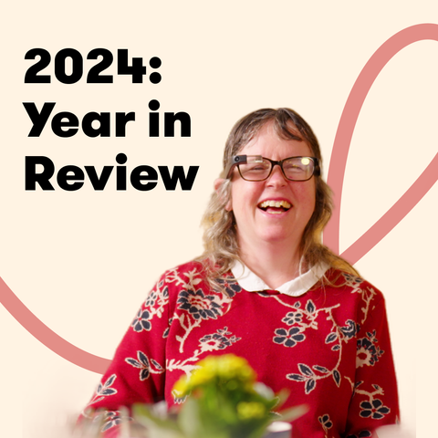 A smiling woman wearing Envision Glasses and a red floral sweater sits in front of a blurred green plant, with text reading '2024: Year in Review' on a light beige background with a red looping design