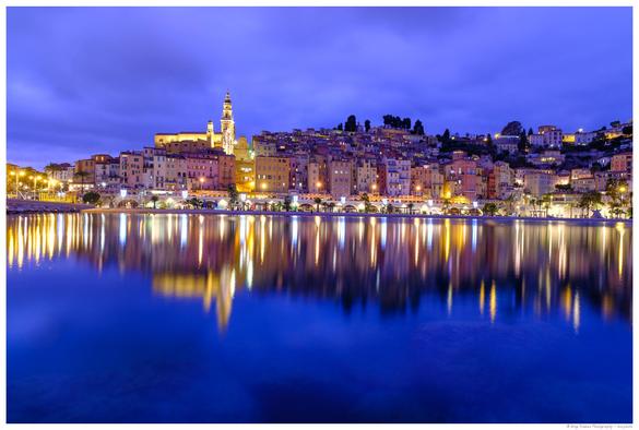 A blue hour shot of a French old town on a hill on the seaside, with a well-lit church on the left-hand half of the image. The sky is cloudy and smooth and the sea looks smooth due to a long exposure, reflecting the city hill above it. Everything but the yellow city lights is deep blue. 
