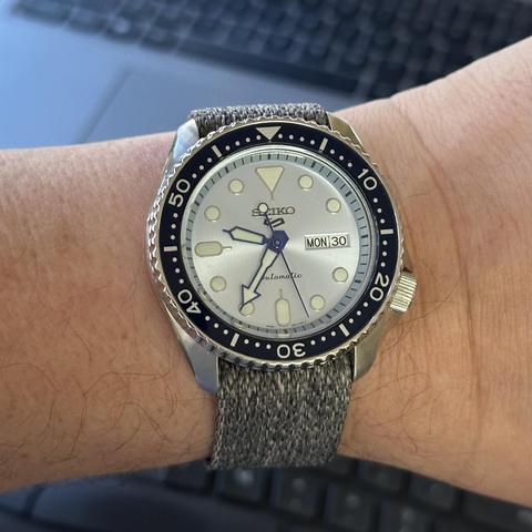 Watch with stainless steel case with brushed upper surfaces and polished sides, a 4:00 crown, an ice blue dial with day/date at 3:00, blue diver-style arrow hands and blue seconds hand, diver-style applied indices, and gray tweed-like fabric strap. 