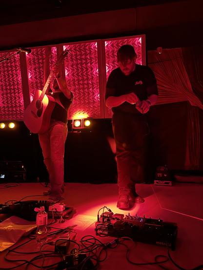 the band pornophonique live on stage in the lounge at 38c3. kai holding his guitar up in the air, felix is turning off the gameboy. lights are red. the shot was probably taken at the ending of a song or maybe the whole set.